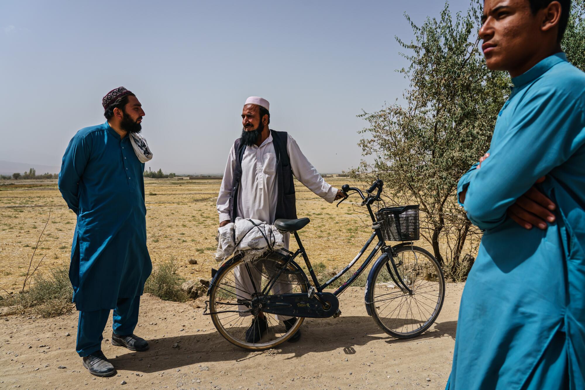 A man with a bicycle talks to two others on a rural road with an empty field in the background.