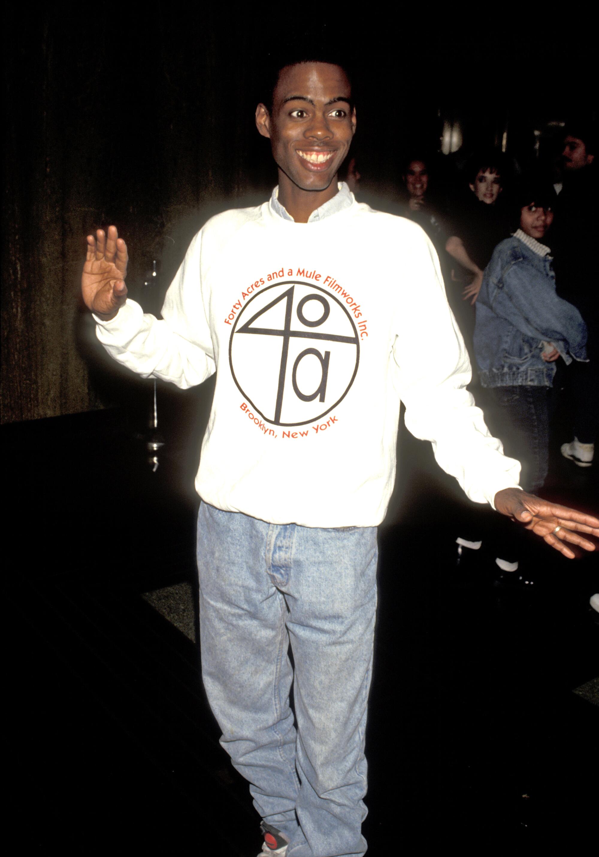 Chris Rock in a white sweatshirt and jeans with arms partially outstretched.