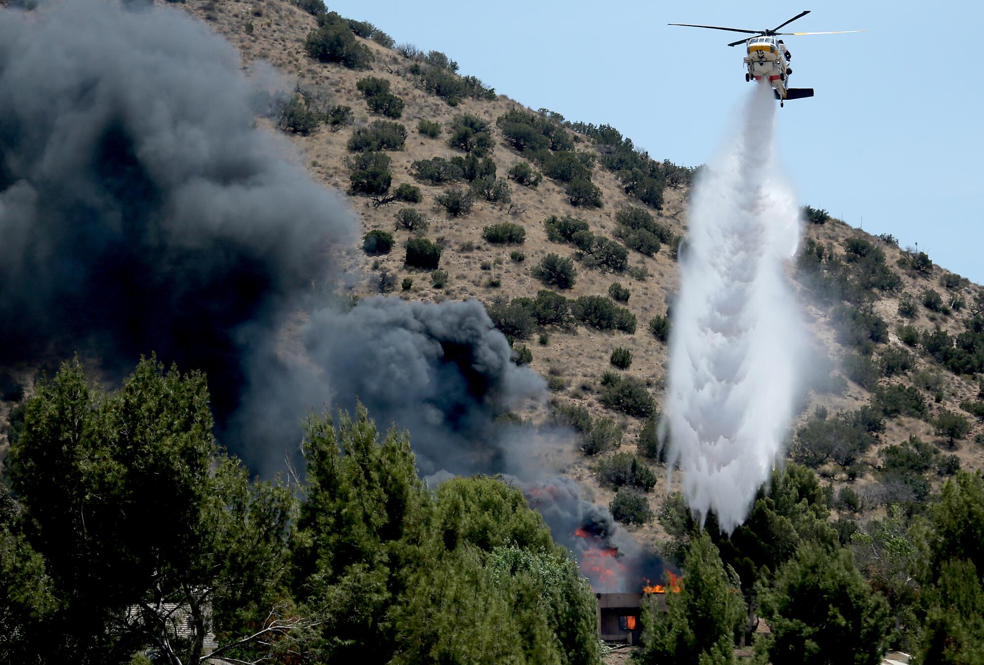 Above a scrub-covered hillside a helicopter hovers and releases a white plume on a burning structure.