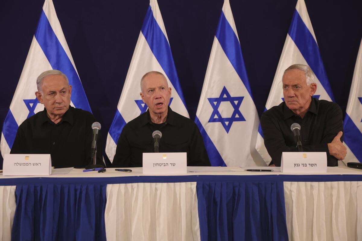 Three men in dark clothing seated at a table with microphones and blue-and-white flags behind them 