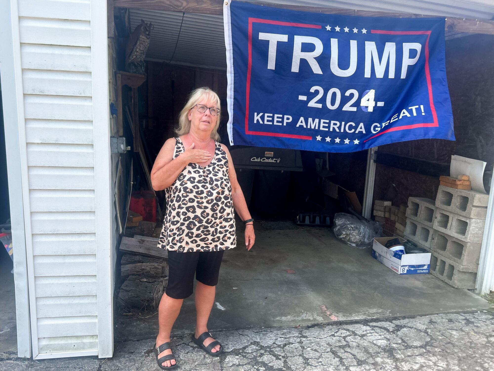 A woman stands in front of a garage with a "Trump 2024" banner.