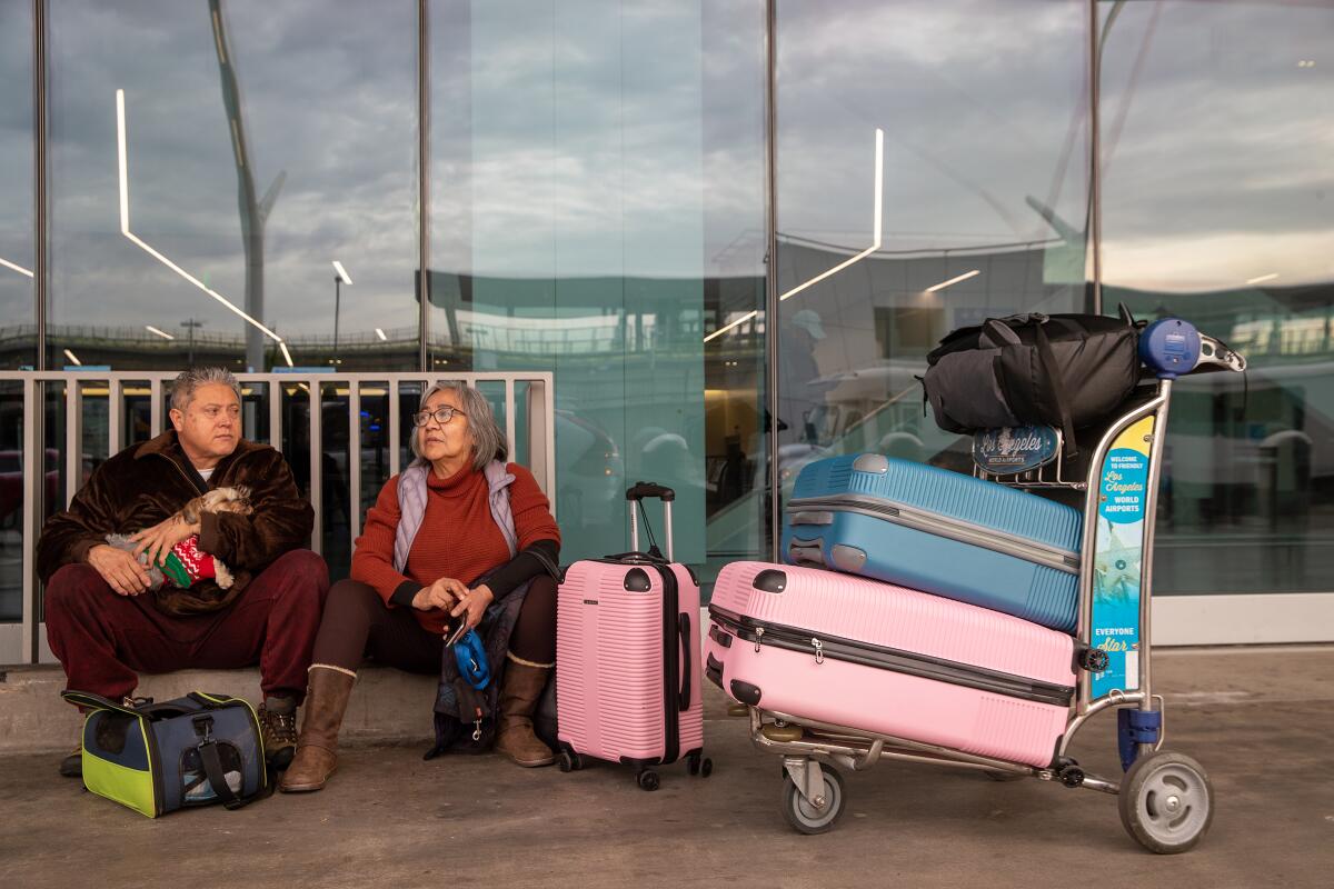 Two people, one holding a small dog, sit next to a cart loaded with suitcases outside of an airport building. 