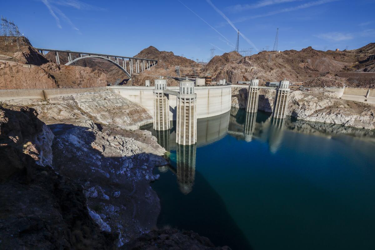 A rocky shore, a lake and a dam are seen. In the background a bridge spans a canyon.