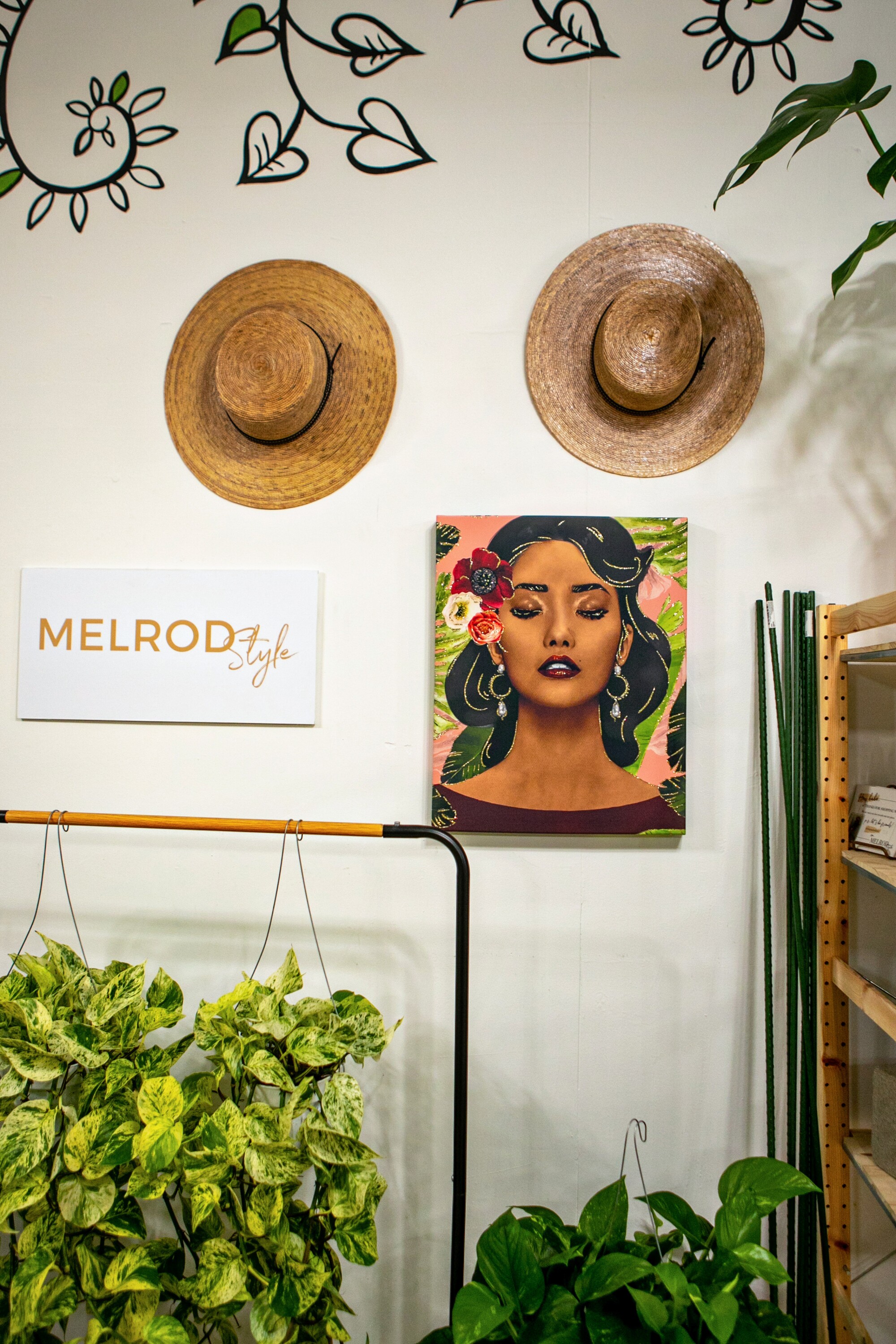 Two hats and a painting of a woman hang on a wall above some potted plants. 