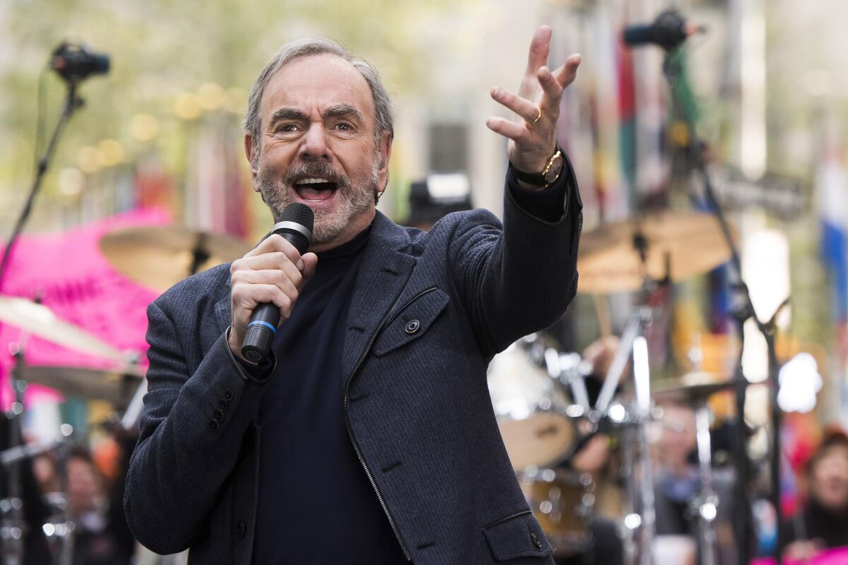 Neil Diamond holds a microphone in one hand and gestures with his other hand onstage