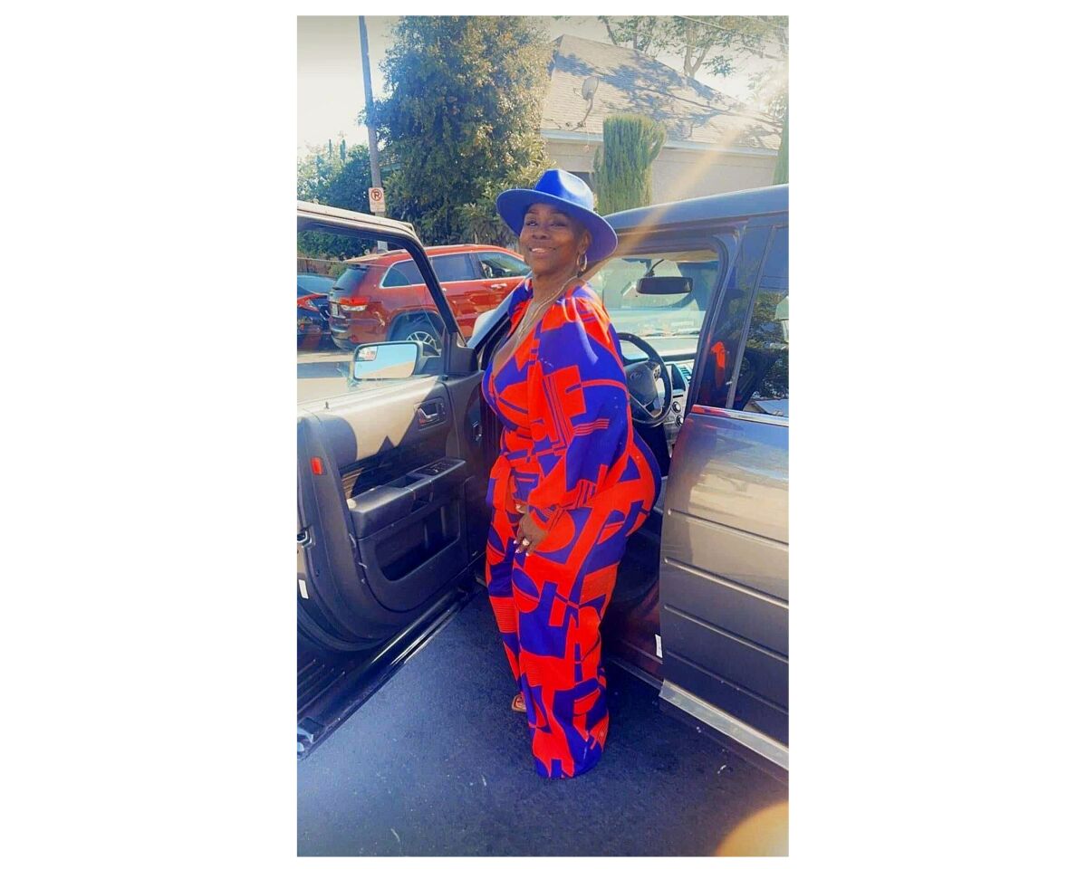 A woman stands next to the open door of a car wearing a blue brimmed hat and a flowing pantsuit.