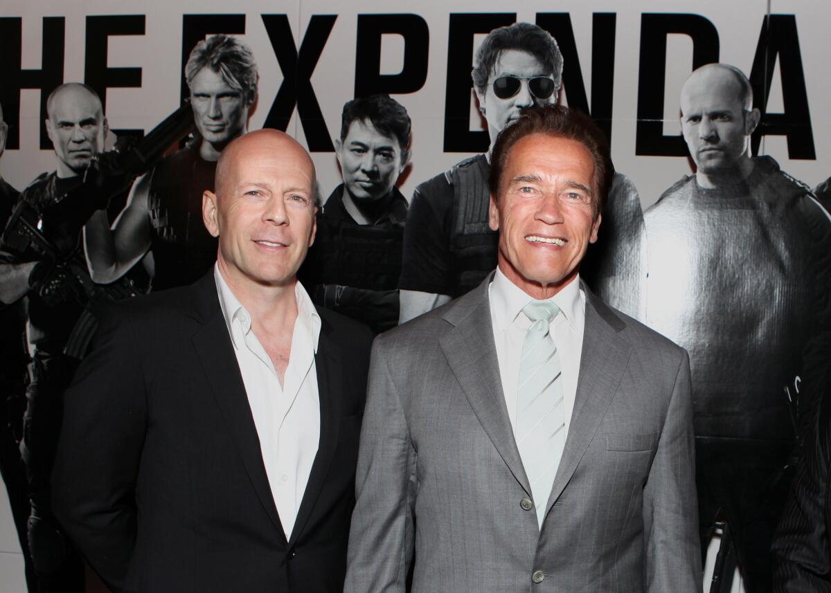 Bruce Willis in a suit shirt and black blazer standing next to Arnold Schwarzenegger in a gray suit. Both men are smiling