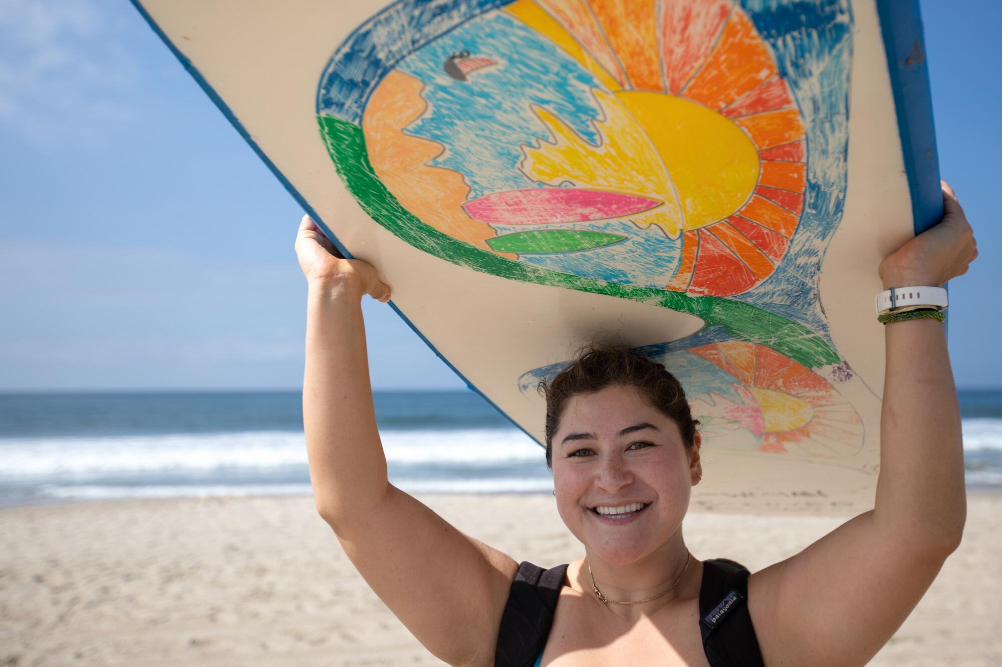 A woman on the beach holds a surfboard over her head.