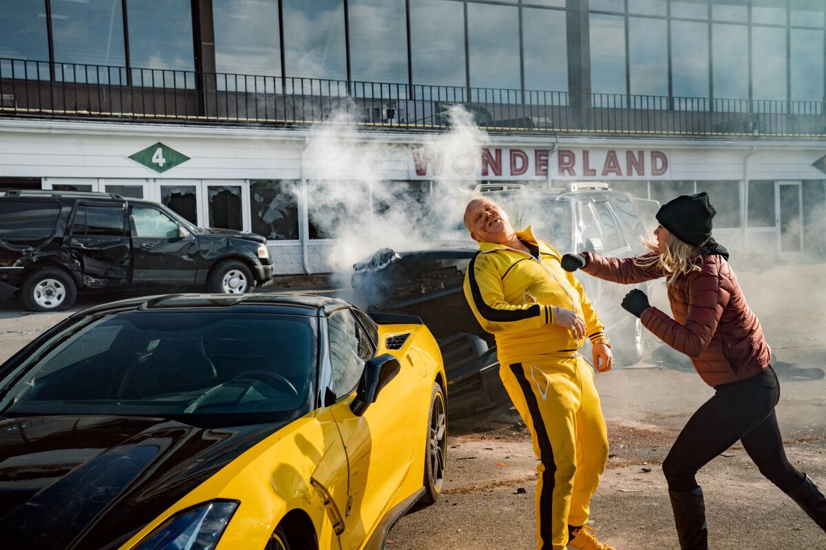 Iliza Shlesinger throws a punch at James DuMont, with a yellow tracksuit and sports car, in "Spenser."