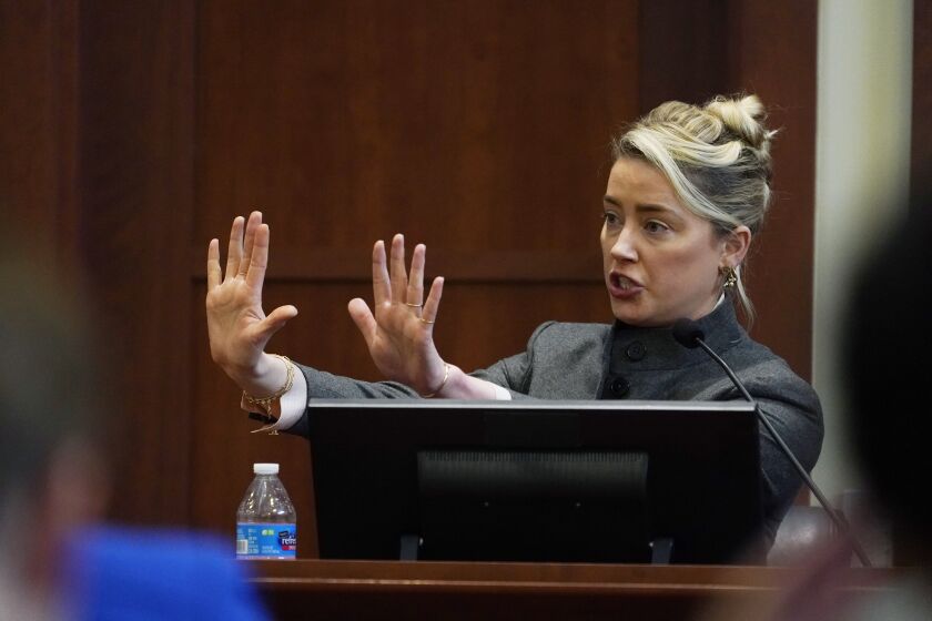 A woman on a witness stand raises two hands in front of her in a warding-off gesture