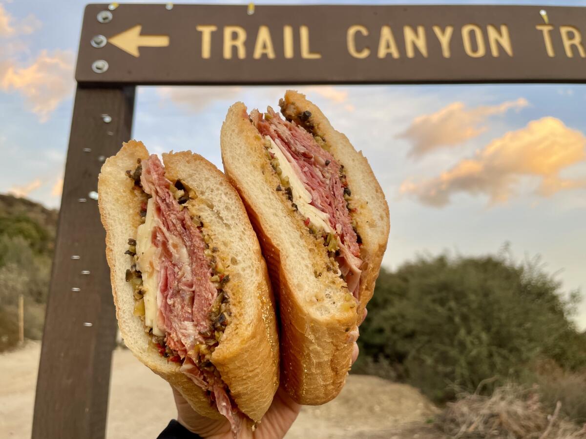 A hand holds up two halves of a sandwich in front of a trail sign.