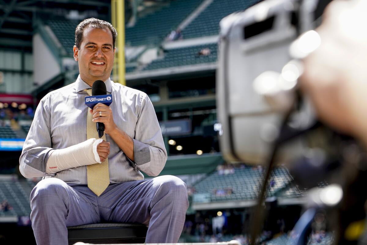 A man with his right arm in a cast holds a microphone with his left hand while sitting in a stadium.