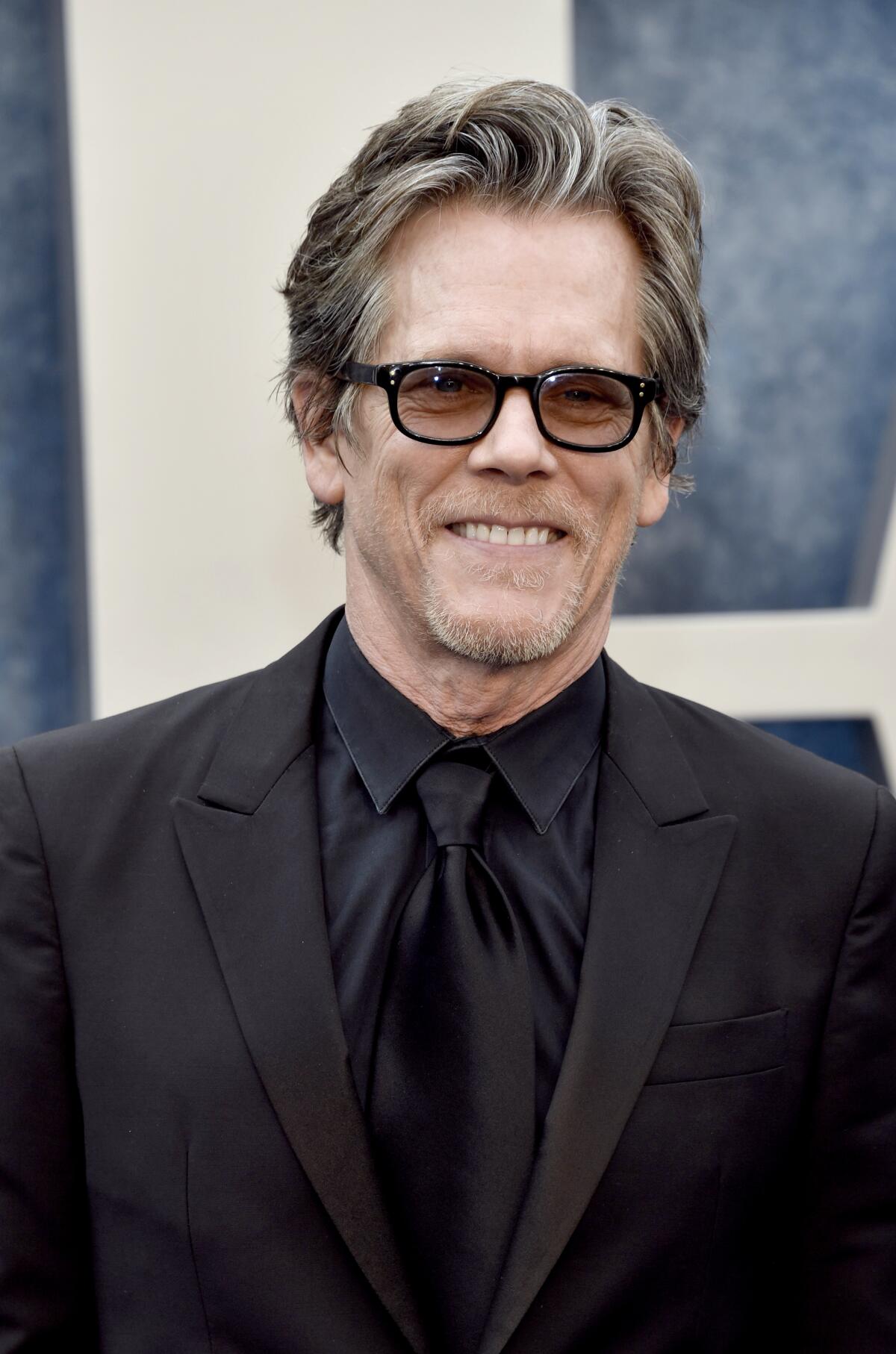 Kevin Bacon smiles while posing in dark glasses and a black suit and tie