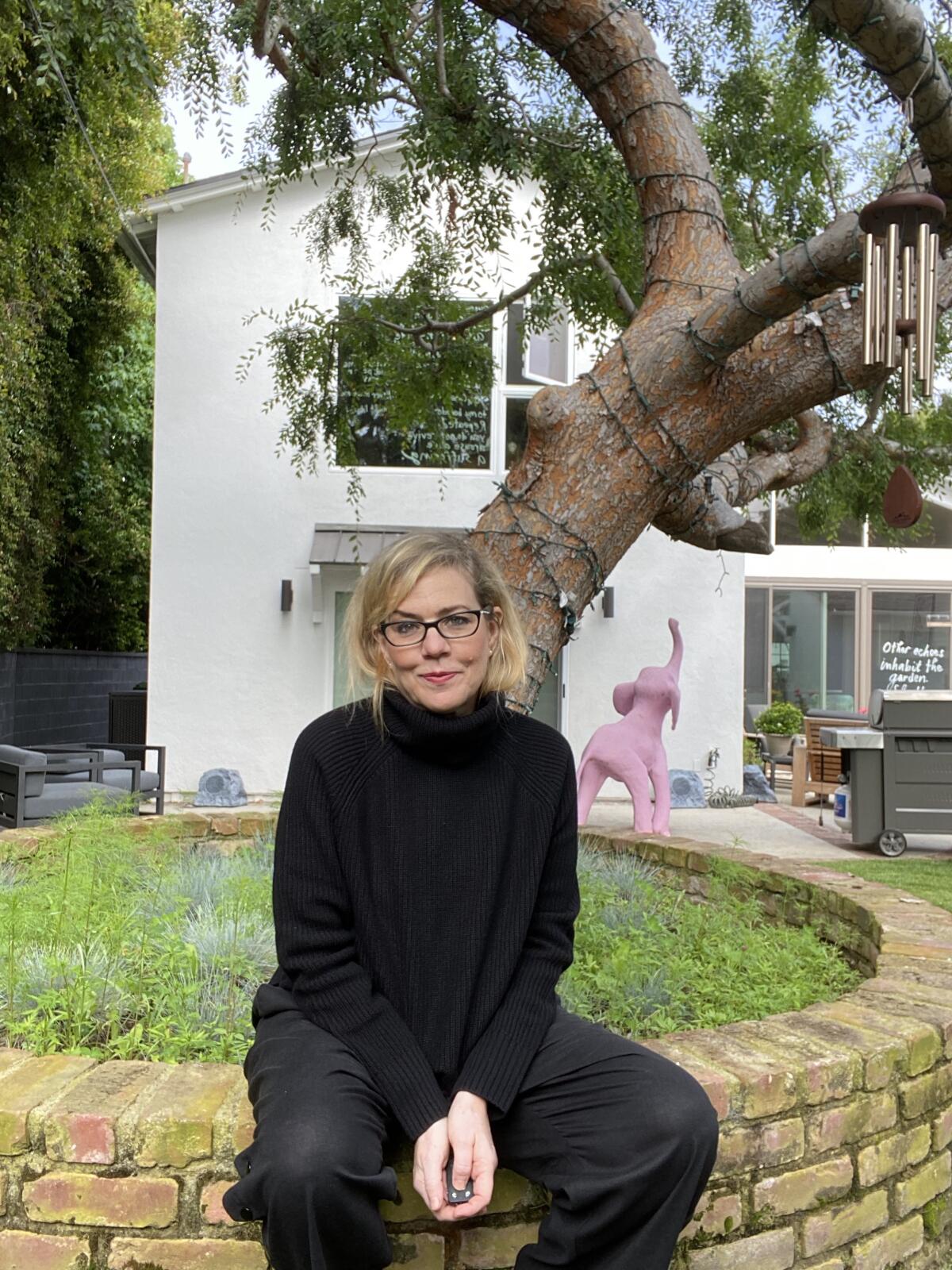 A woman in a black turtleneck sits on a raised brick garden bed 