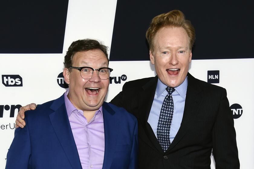 Andy Richter and Conan O'Brien pose together in suits while smiling with their mouths open.