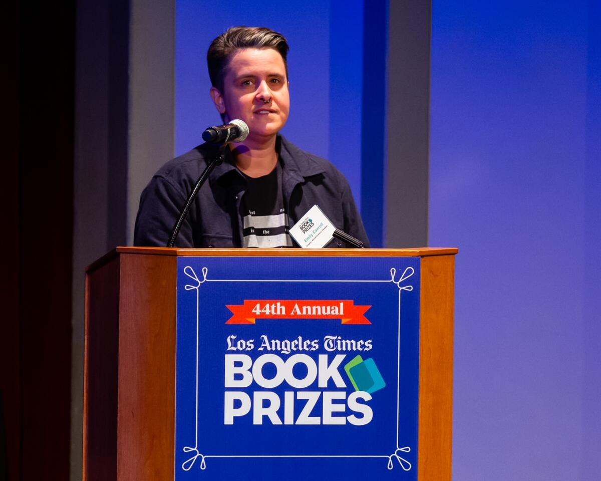 A woman in a black T-shirt and black jacket stands at a lectern with the Los Angeles Times Book Prizes logo.