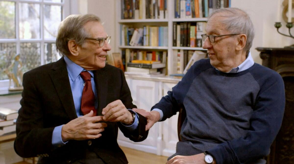 Two older men in a book-filled office smile and talk to each other