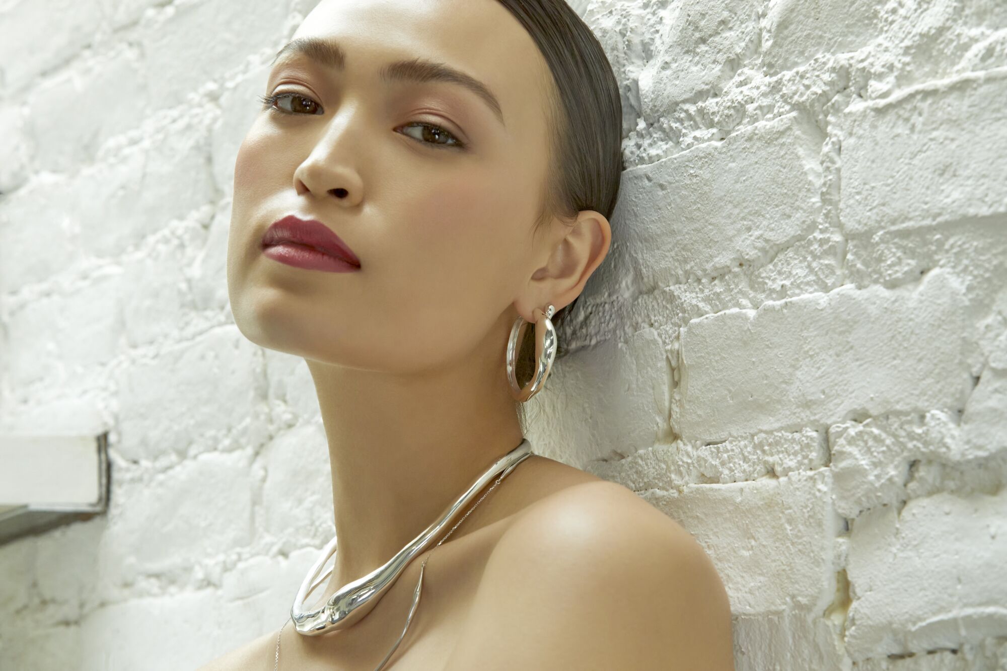 A woman wearing silver jewelry leans her head against a white brick wall.
