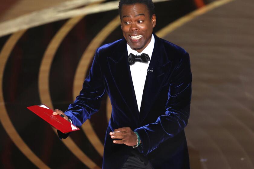 Chris Rock, holding a red envelope and wearing formal attire, speaks onstage during the Oscars