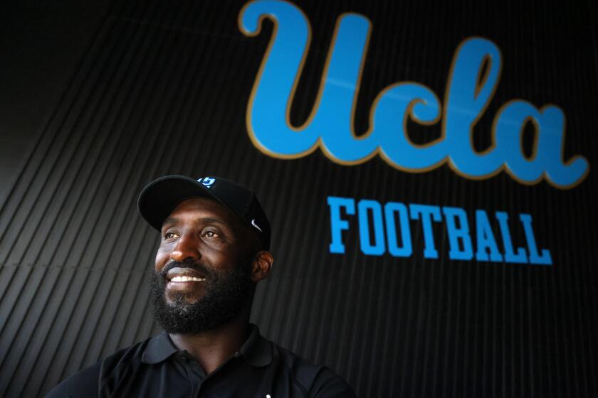 UCLA football head coach DeShaun Foster smiles while standing near a "UCLA football" sign inside the practice facility 