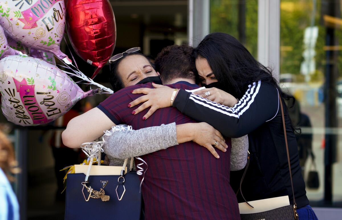 Three people share a group hug alongside balloons that say "Best Mom ever!"