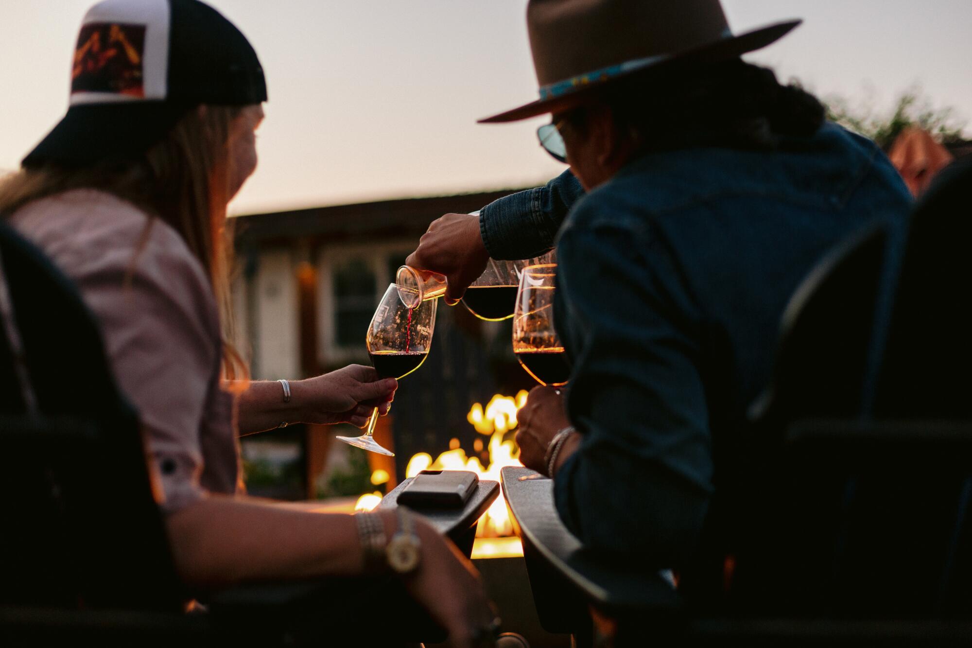 A view from behind of a man pouring a glass of wine for a woman while seated around a bonfire.