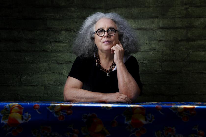 Alison Saar, wearing a black top and a chunky necklace, poses in dramatic light behind a table with a bright cloth