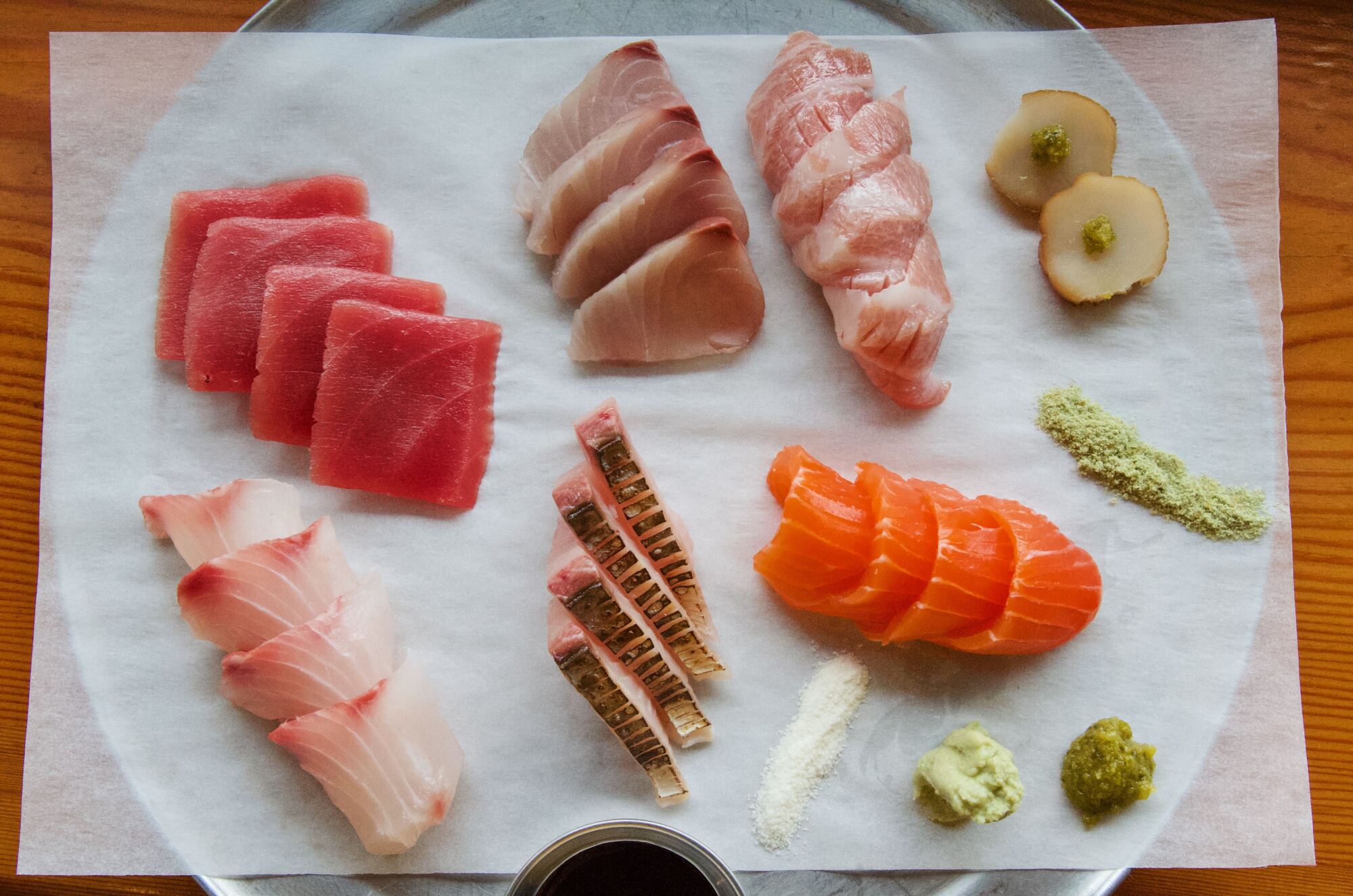 An overhead photo of cuts of a variety of fish on a silver platter: trout, tuna, tuna belly and more.