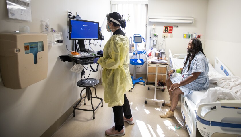 زنی با ماسک و محافظ صورت جلوی صفحه کامپیوتر ایستاده است.  پشت سرش مردی بود که روی تخت بیمارستان نشسته بود