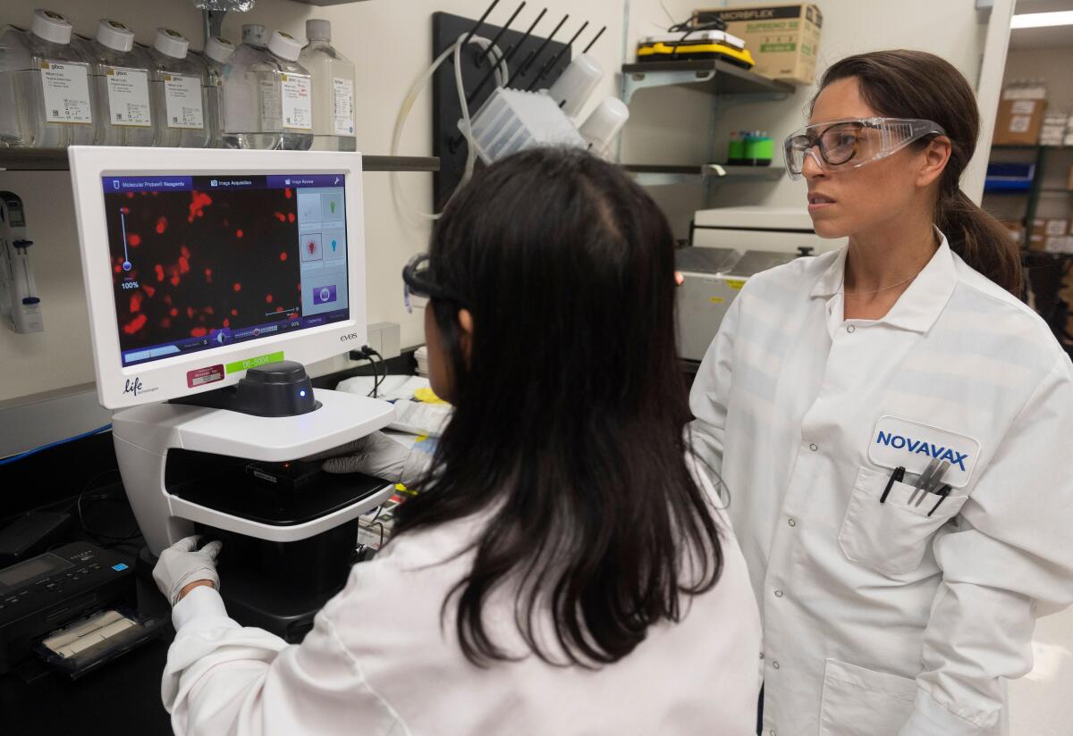 Two women in white lab coats, plastic gloves and goggles look at a computer screen.