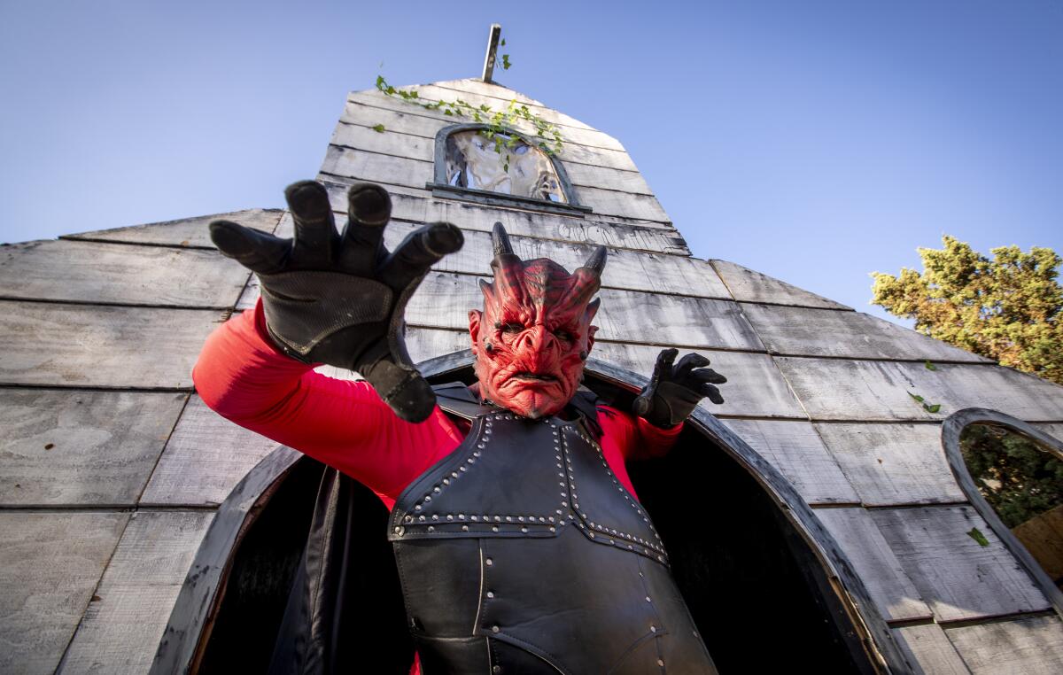 A person in a devil costume stands in front of a building made of gray boards.