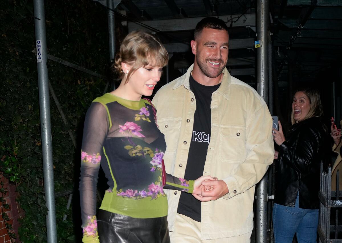 Taylor Swift in a sheer top and black skirt holds hands with Travis Kelce in a beige jacket-pant set and black shirt.