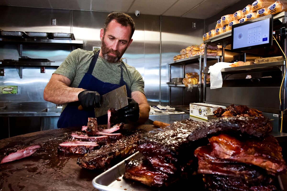 A bearded man in a T-shirt and chef's apron cuts up barbecued ribs in a professional kitchen.