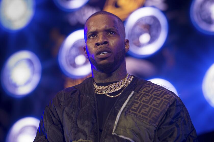 A rapper in a dark jacket and multiple gold chains looks slightly upward while onstage