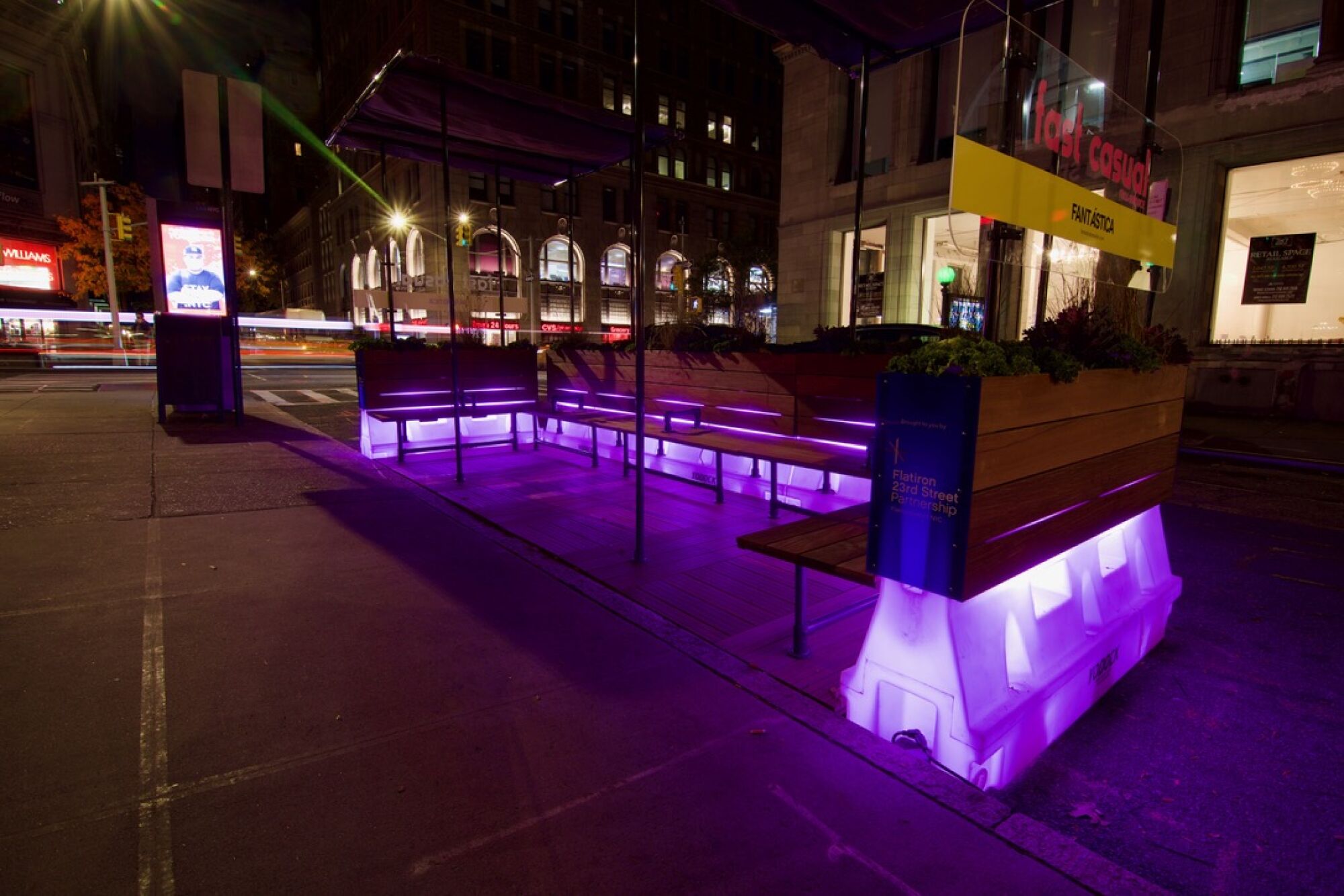 A parklet designed with illuminated lavender-colored water barriers is seen at night