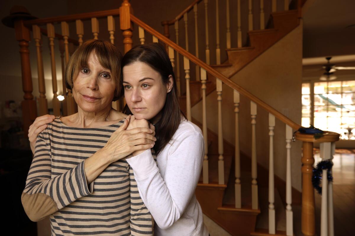 A mother and her adult daughter embrace in front a staircase in their home. 
