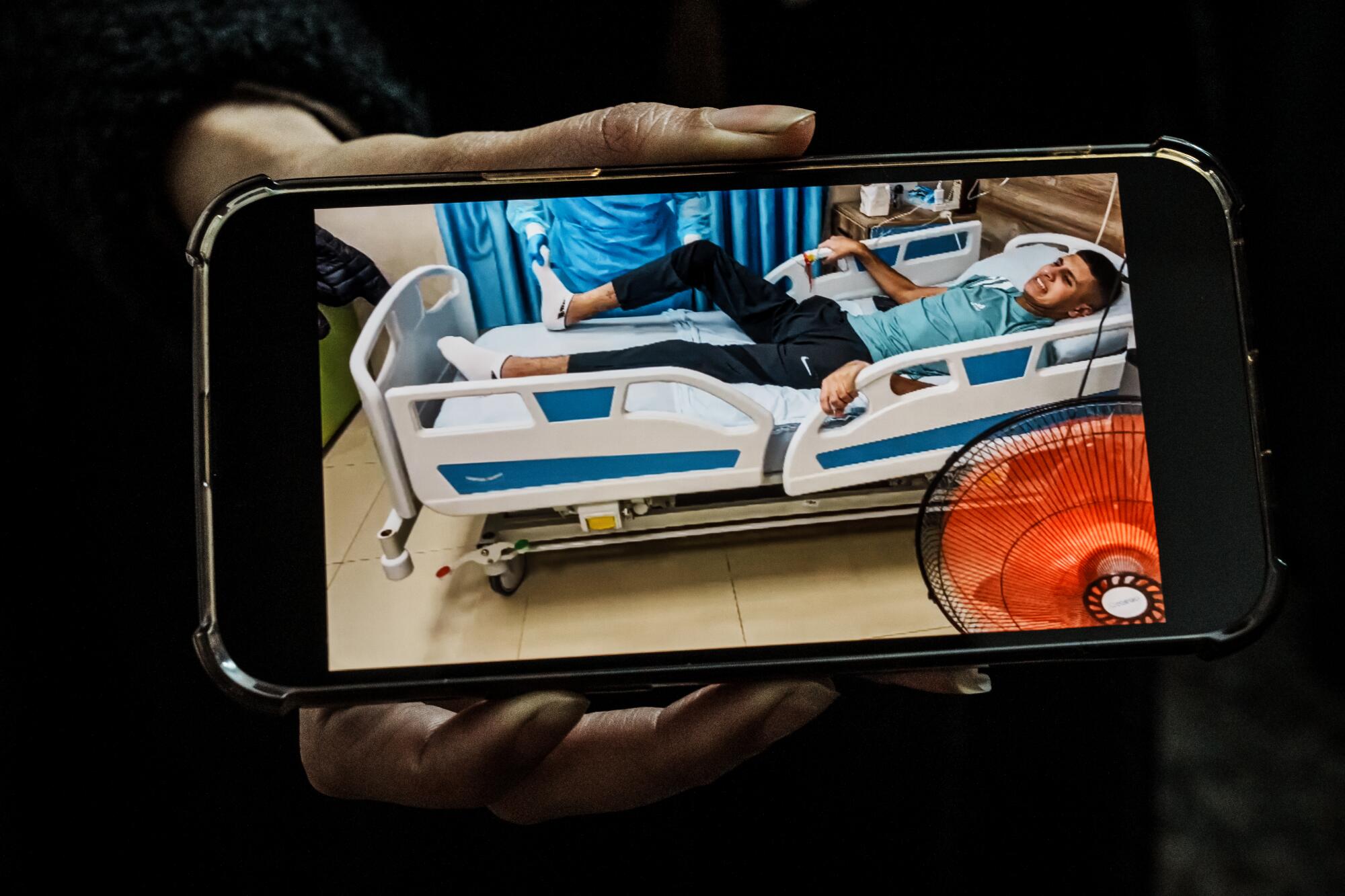 An image on a phone shows a man lying in a hospital bed with one leg partly raised 