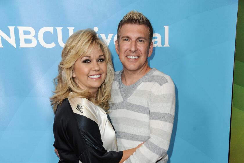 A blonde woman in a blue top and white pants embraces a blond man in a grey striped sweater and pink pants