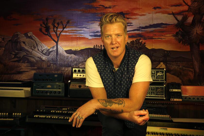 Josh Homme wears a white t-shirt and black vest as he leans on a soundboard in a studio