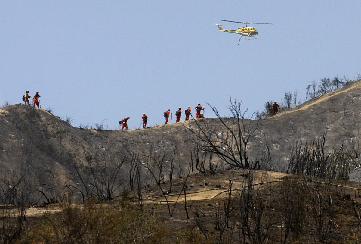 As a water-dropping helicopter looks for hotspots, a hand crew moves across a burned mountainside Monday above Azusa.