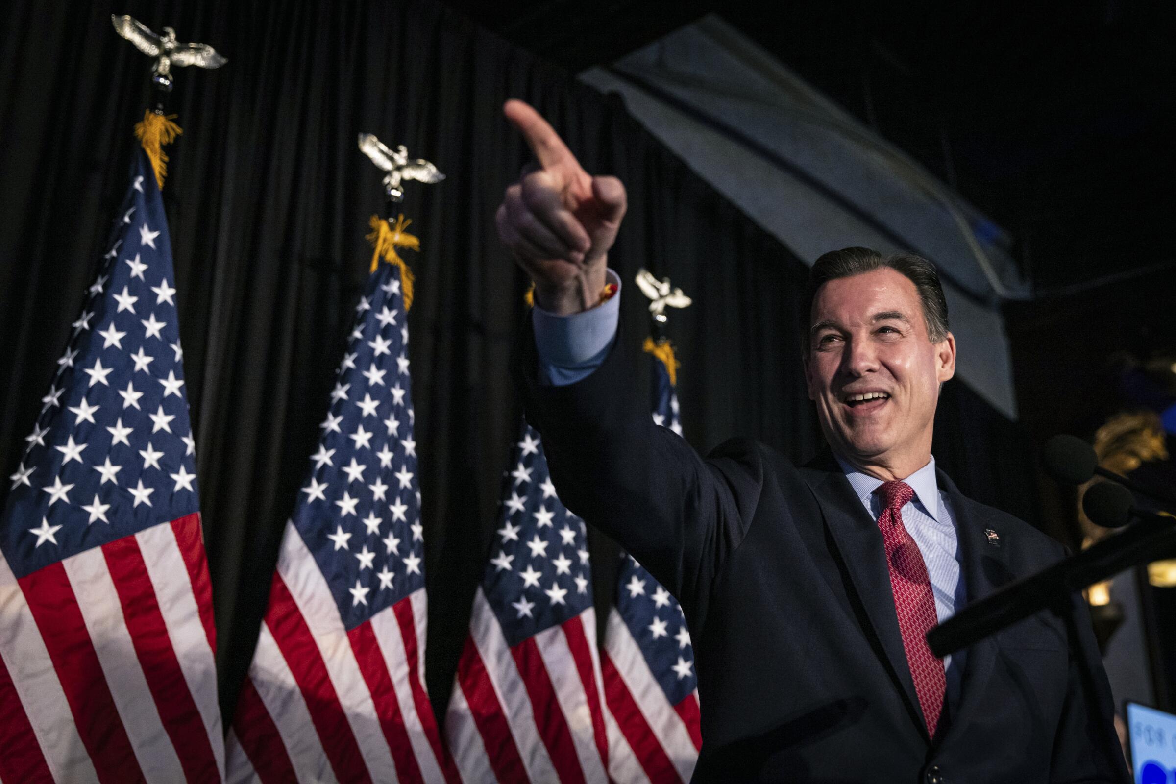 Tom Suozzi points at supporters from a stage in front of four furled American flags 