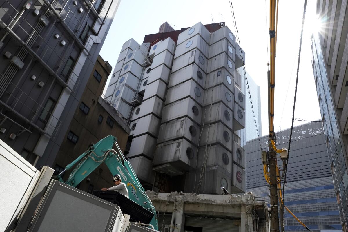 A crane and members of a construction crew are seen next to the Nakagin Capsule Tower, which is wrapped in construction mesh.