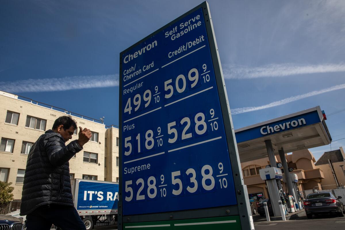 A man stands near a sign displaying fuel prices at a gas station.