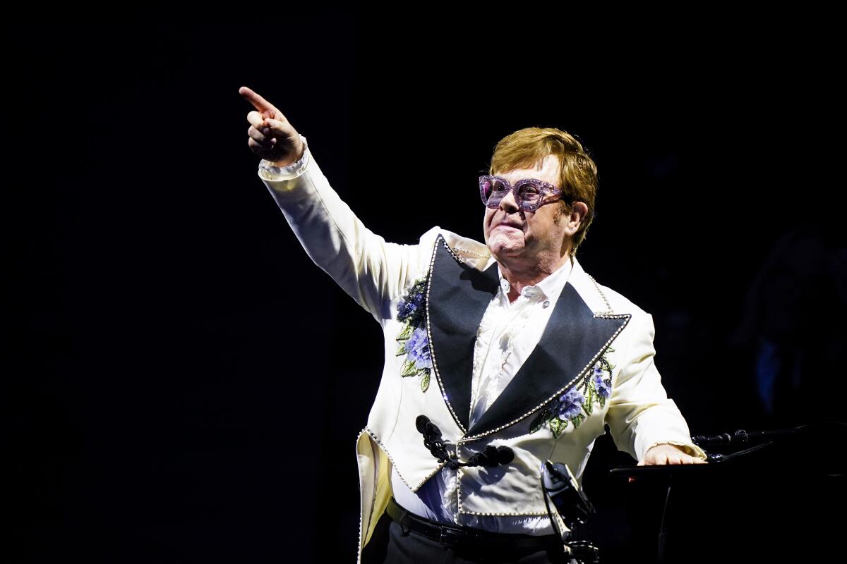 A man in a white suit and sparkly glasses sits at a piano and points upward.