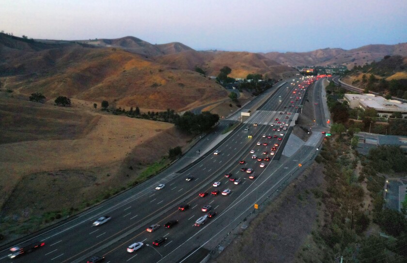 An aerial view of a freeway filled with cars and running past low brown hills.