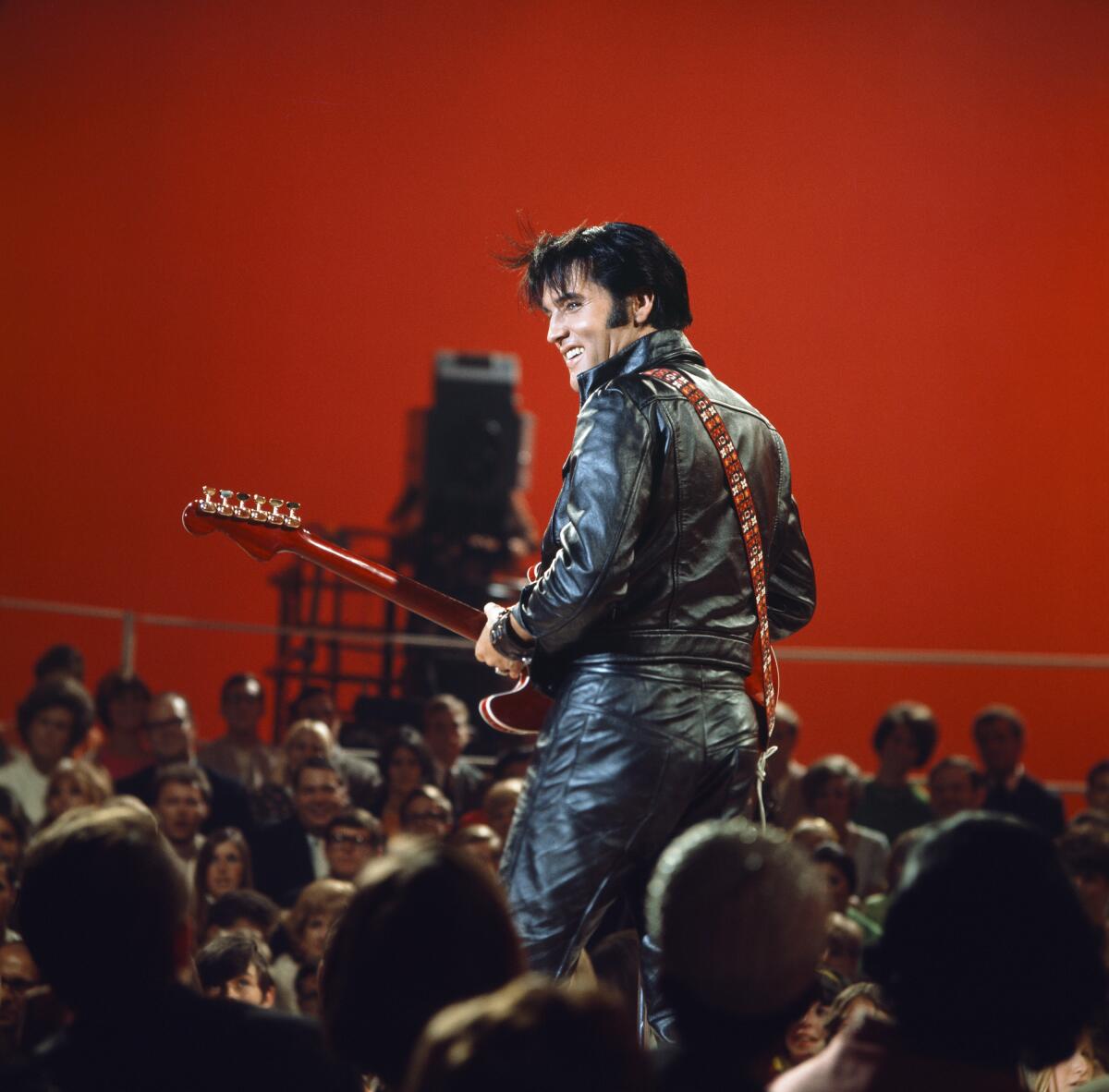 Elvis Presley from behind, surrounded by an audience. He is in a black leather suit and is holding a guitar.