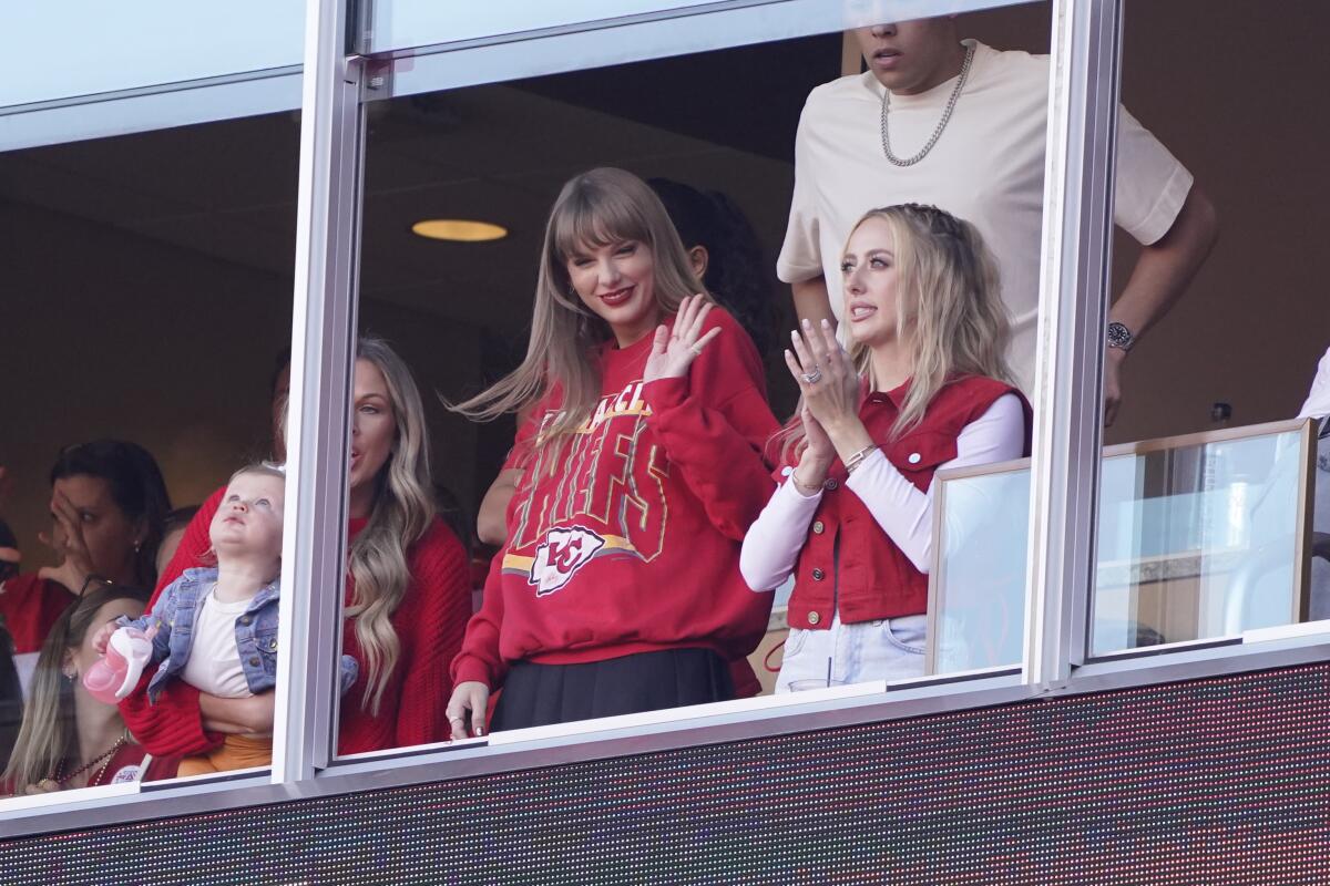 Taylor Swift wears a Kansas City Chiefs sweatshirt and waves from a luxury box at a football game.