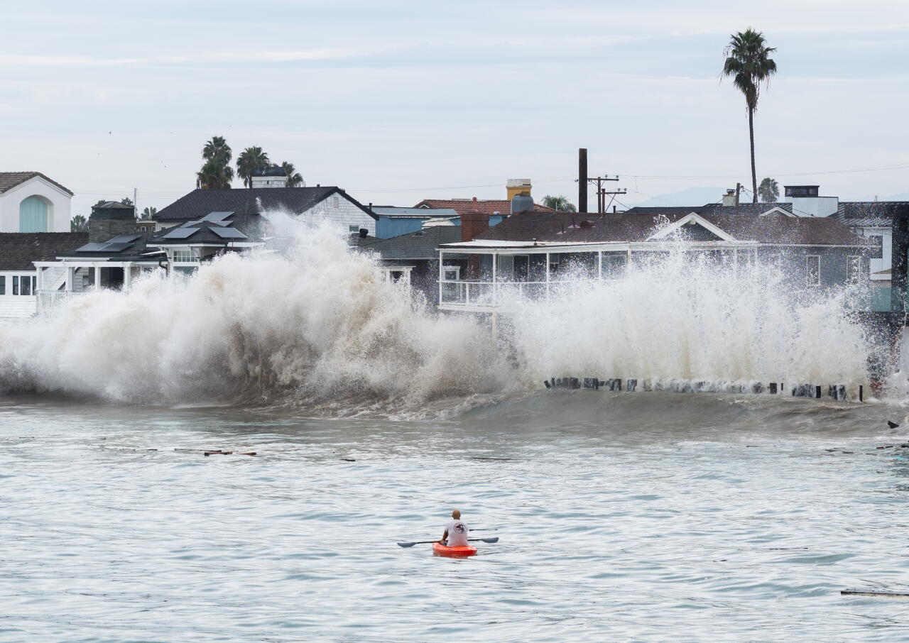 A person in a kayak is in the foreground looking at homes and a wall of waves.