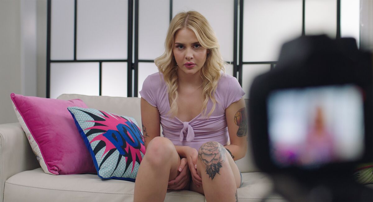 A woman with tattoos sits on a couch next to some throw pillows in the 2021 drama “Pleasure.”