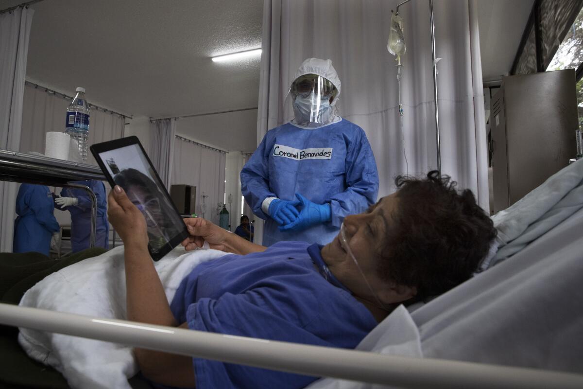 A patient speaks to her daughter on a tablet while in bed at a hospital while a hospital staff member stands nearby.